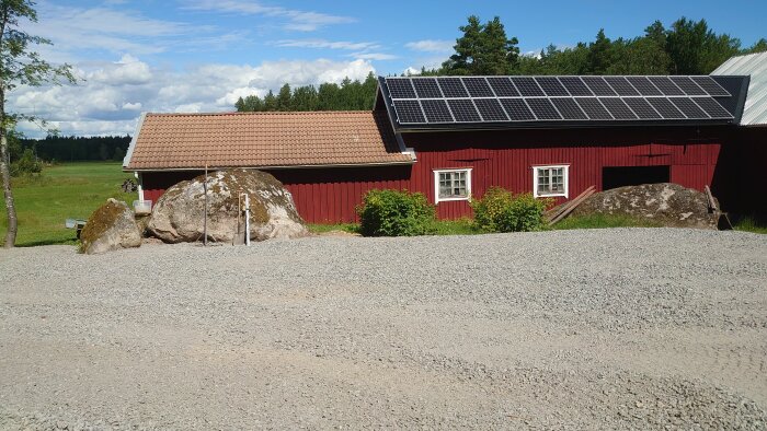 Traditionellt rött trähus med solpaneler på taket, stora stenar, grusplan, och grönskande landskap i bakgrunden.