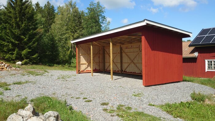 En röd träbyggnad, troligen ett förråd eller vedbod, med grusgrund framför och gröna träd i bakgrunden.