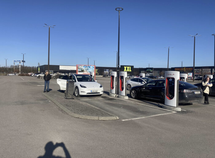 Tesla-laddstation på parkeringsplats med bilar och människor en solig dag.