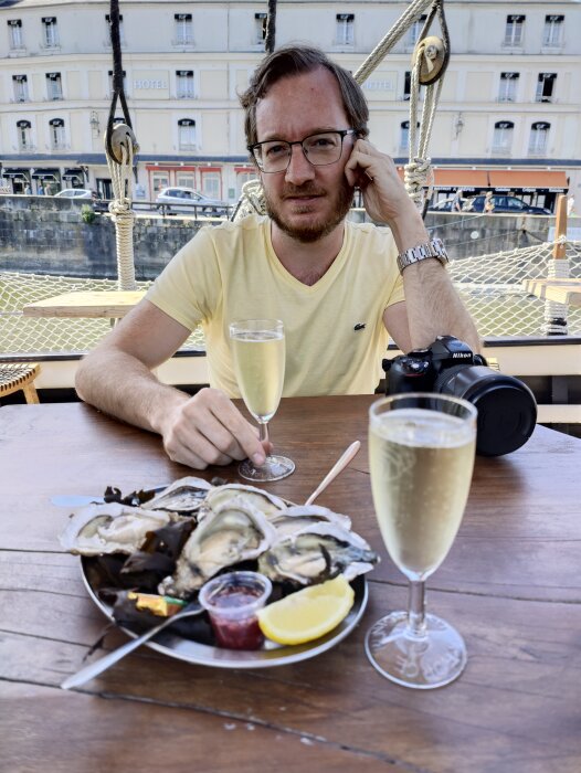 Man äter ostron och dricker champagne på en utomhusrestaurang med hamnutsikt. Kamera på bordet.