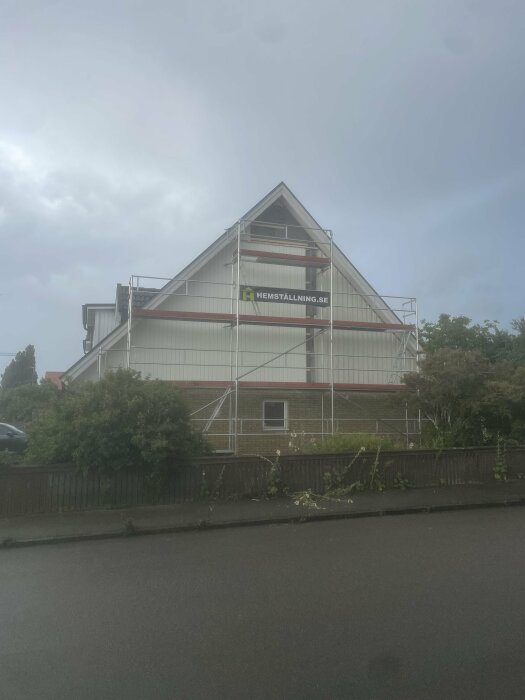 Ställning runt huset under renovering eller målning, molnig himmel, grönska, villakvarter, reklamskylt.