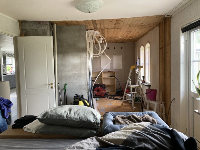 Inre rum under renovering med säng, byggmaterial, verktyg och stege. Oavslutat arbete, rörigt, inhyst tillfälligt sovrum.