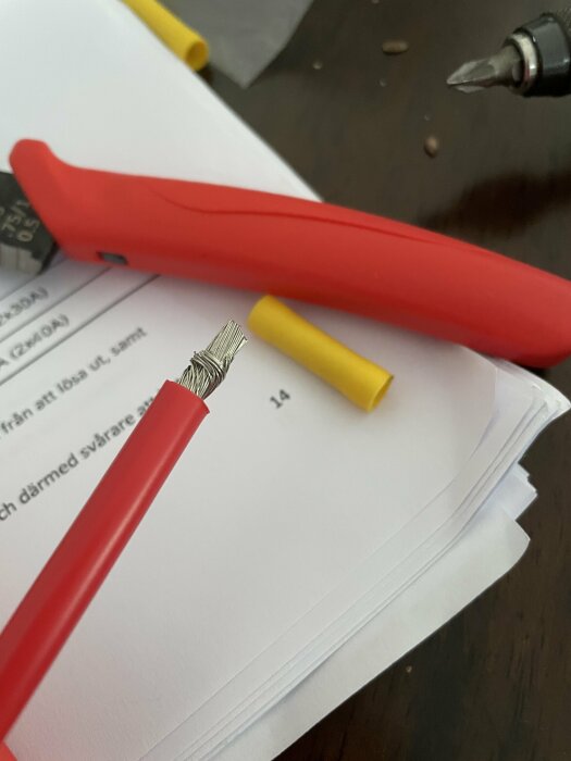 Rödpennans huvud, papper, silvergrå spiraler, gult fodral, träbord, oskarpt skruvmejselspets i bakgrunden.