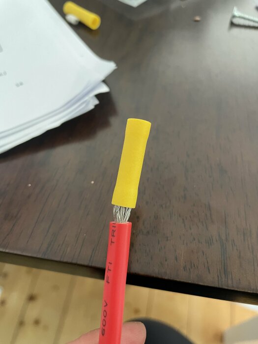 Ett rött verktyg med en gul ändhylsa och synliga metallfibrer, hålls mot en suddig bakgrund med dokument.