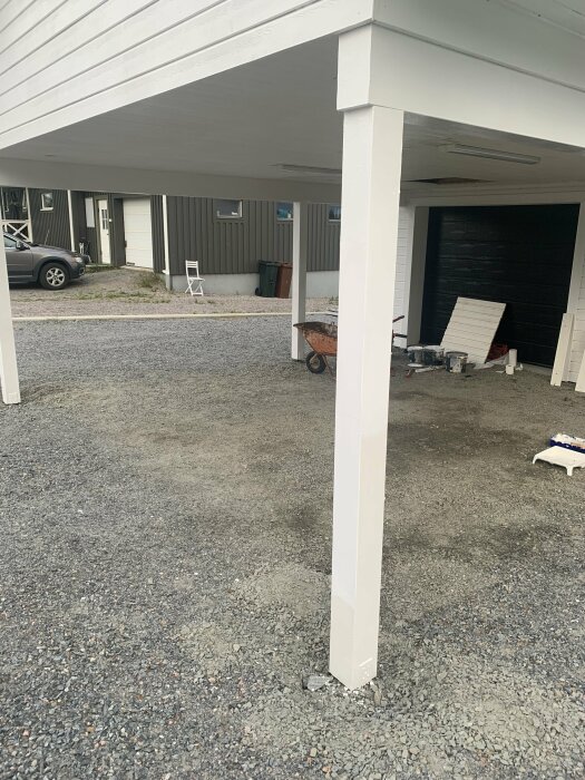 Carport med vit pelare, skottkärra, hus i bakgrunden, grusgolv, målningstillbehör synliga.