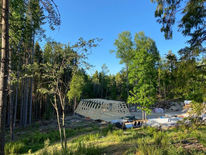 Hustakstomme under uppbyggnad i skogsmiljö, byggmaterial utspritt, klart väder.