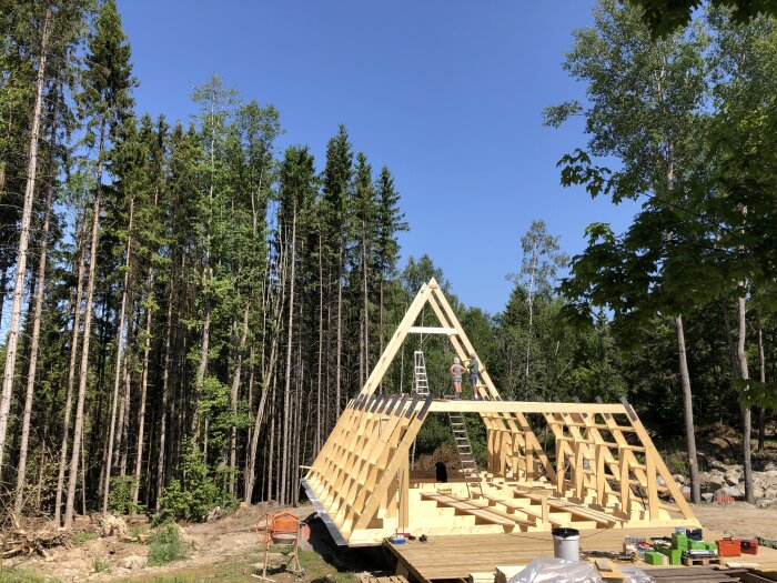 Träkonstruktion av hus under uppbyggnad, arbetare på stege, klarblå himmel, skog i bakgrunden.