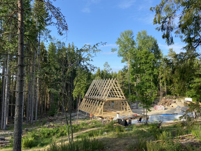 Byggarbetsplats i skogen med trästomme för hus, byggmaterial, och träd runt omkring. Soligt och grönt landskap.