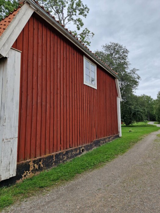 Röd träbyggnad, vit knut, fönster, skadad nederdel, grusväg, grönt gräs, träd, molnig himmel.