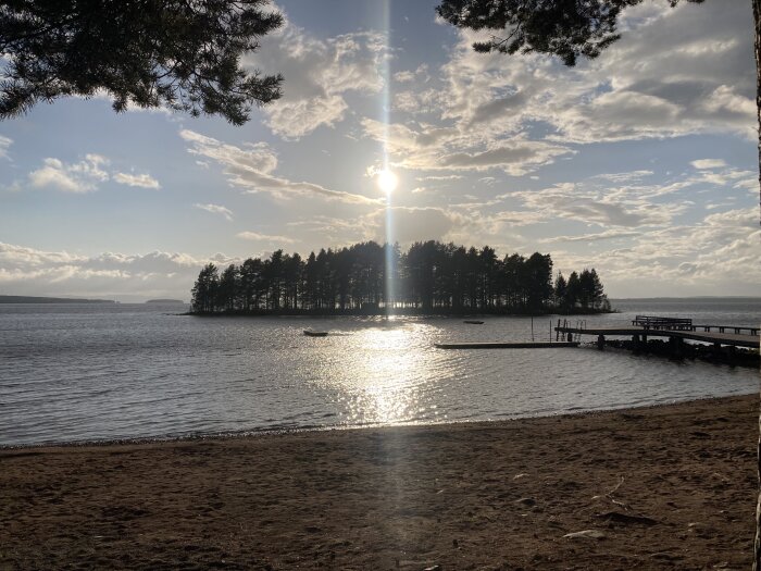 Solnedgång över en sjö, silhuett av en skogsbeklädd ö, strand, brygga, lugnt vatten, reflekterande solljus.