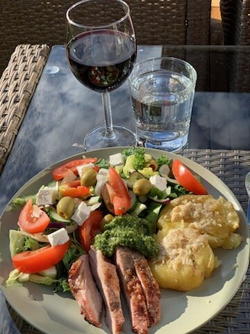 Utomhusmåltid med sallad, grillat kött, potatis, rödvin och vatten, serveras på en solig terrass.