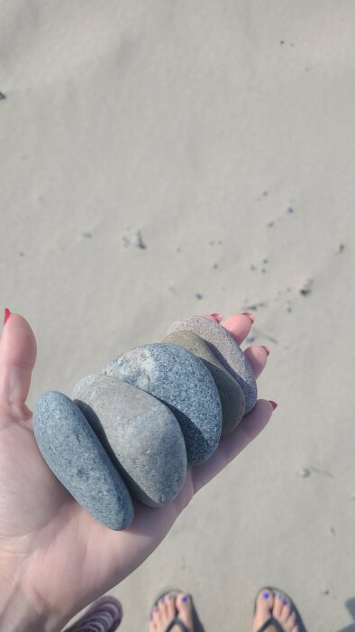 En hand som håller släta stenar på en sandstrand, med fötter och flip-flops i bakgrunden.