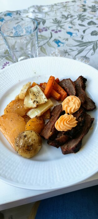 Grillat kött med rostade rotfrukter och kryddsmör på vit tallrik, vattenglas, blommigt bordstyg.