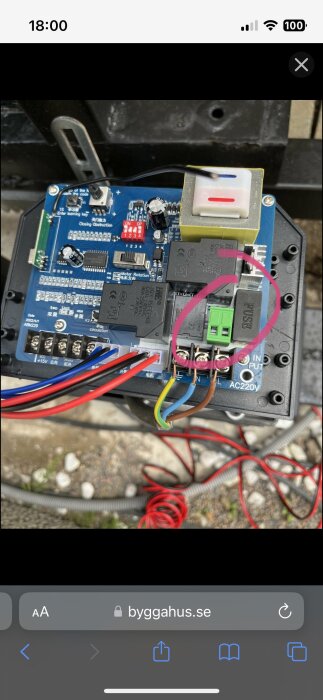 Elektronisk kretskort med anslutna kablar och komponenter, öppet kapsling, rosa markerad del, grusig bakgrund.