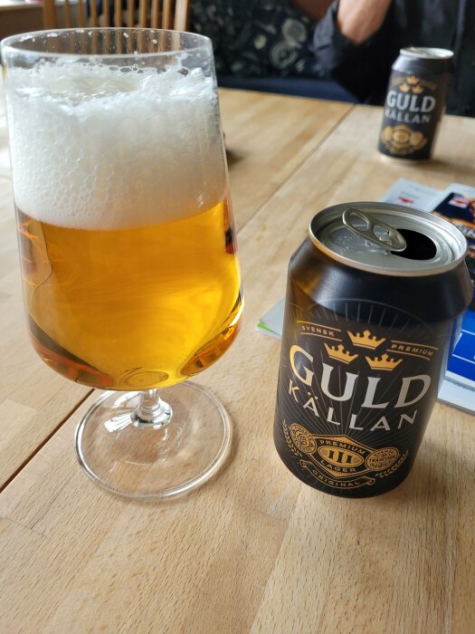 Ölglas med skum, burk öl märkt "GULD KALLAN", träbord, del av person bakgrund.