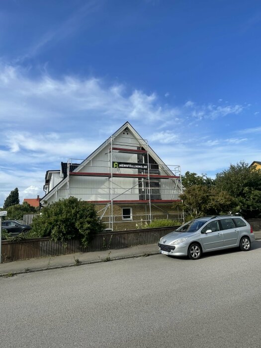 Hus med byggnadsställningar, bil framför, blå himmel med vita moln, grönska, "HEMSTÄLLNING.SE" skylt.