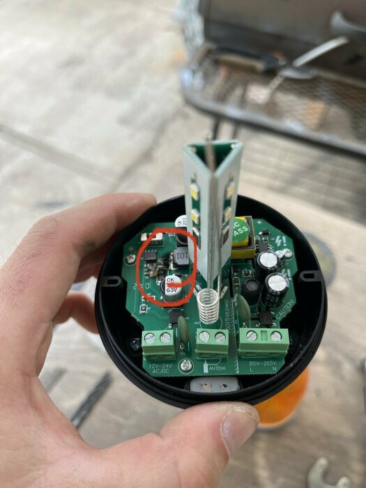 En hand håller en elektronisk enhet öppnad med grönt kretskort, kondensatorer och coil, eventuellt en del av en större apparat.