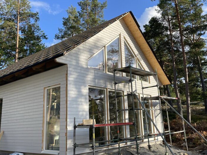 Modernt hus under konstruktion, stora fönster, skogsområde, blå himmel, byggnadsställningar vid fasaden.