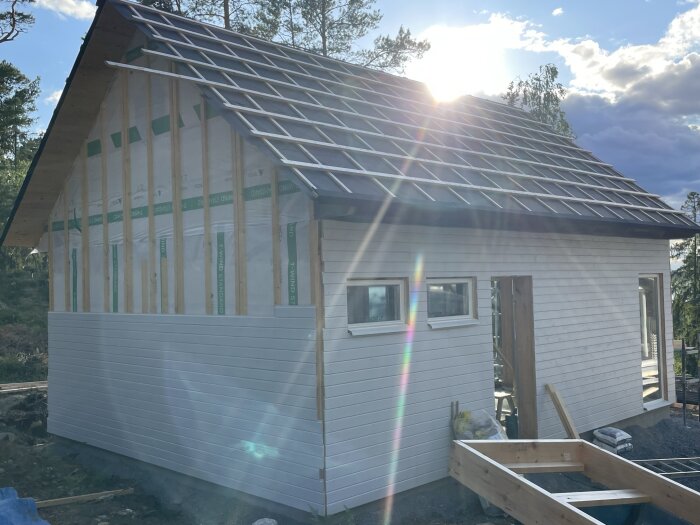 Träkonstruktion hus under uppbyggnad, vit fasad, oisolerade väggar, soligt, klar himmel, delvis täckt tak.