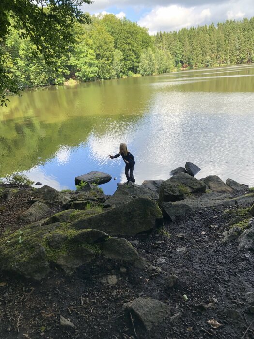 Barn leker vid en skogssjö, klippor i förgrunden, gröna träd reflekteras i vattnet.