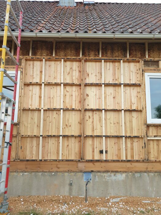 Hus under renovering, träbeklädnad, tegeltak, byggställning, isoleringsmaterial synligt, spill från arbete på marken.