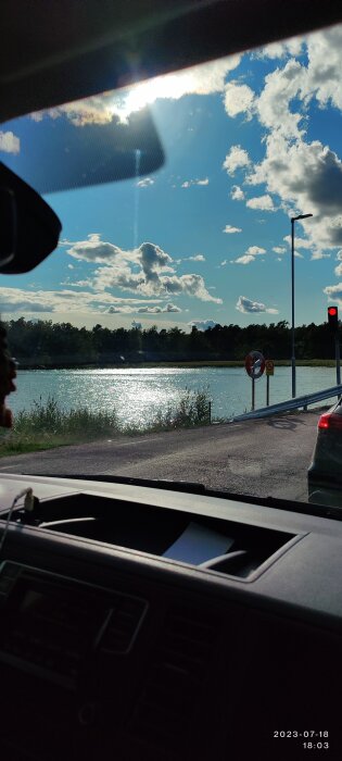 Vy från bil, sjö som glittrar i solen, trafikljus på rött, blå himmel med moln.