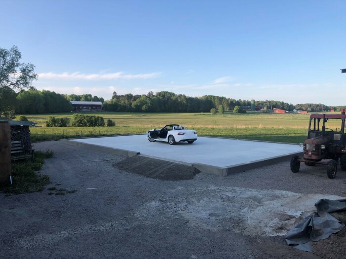 Ny gjuten betongplatta med en vit cabriolet-parkering, lantlig miljö, traktor och byggnadslandskap i bakgrunden.