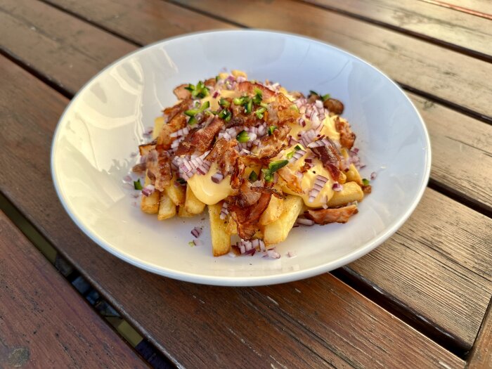Pommes frites med bacon, rödlök, ost, gräslök på tallrik, träbord i bakgrunden.