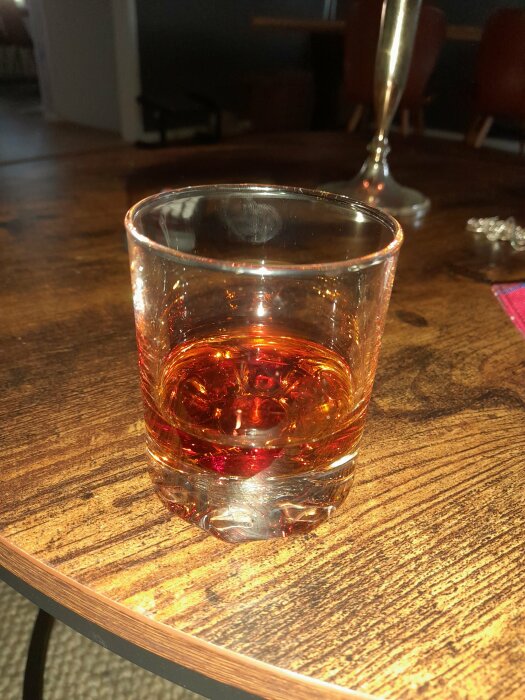 Ett whiskyglas halvfullt med bärnstensfärgad dryck på ett träbord, inomhusmiljö, mysig atmosfär.