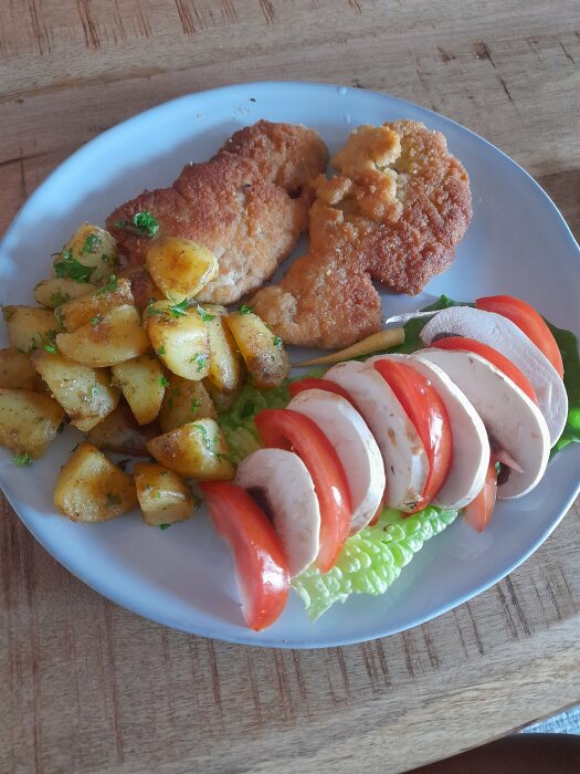 Tallrik med panerad schnitzel, rostade potatisar, sallad med tomat och mozzarella, träbakgrund.