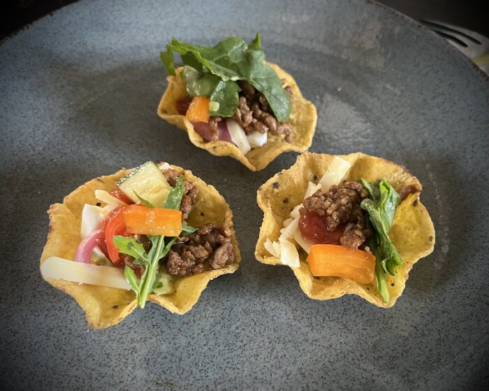 Tre små taco-korgar med köttfärs, grönsaker och ost på en mörk tallrik.
