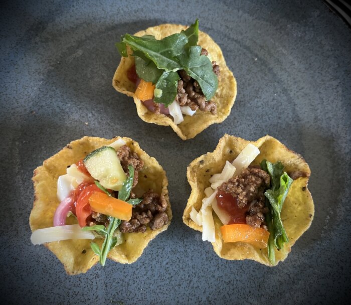 Tre tacos med malet kött, sallad samt grönsaker i små tortillaskålar på en mörk tallrik.