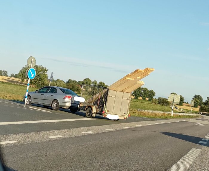 Bil drar släpvagn med överhängande last, risk för osäker transport, på landsväg, nära rondell, klart väder.