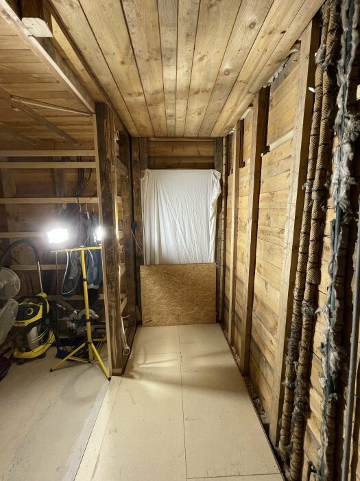 Oisolerat vindsutrymme med träväggar, vit duk, arbetslampor och diverse föremål.
