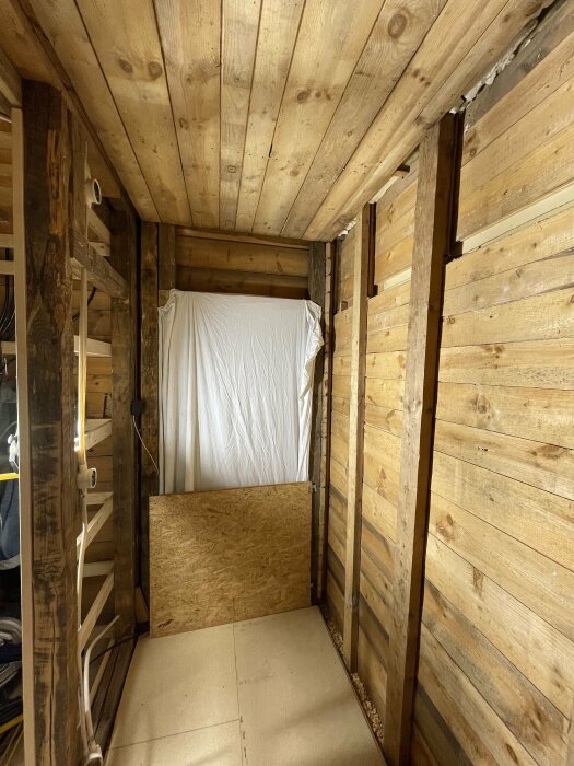 Träbeklätt rum med lakan som dörr, oslipade bjälkar, golv av plywood och betong. Rustik och enkel inredning.