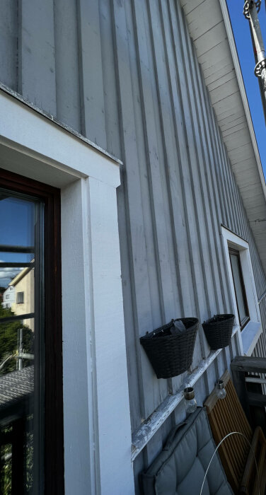 Vertikala träpaneler på fasad, vita fönsterkarmar, svarta korgar, utemöbler, del av balkong.