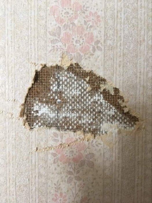 En skadad vägg med delvis bortriven tapet som avslöjar underliggande brunmönstrat lager.