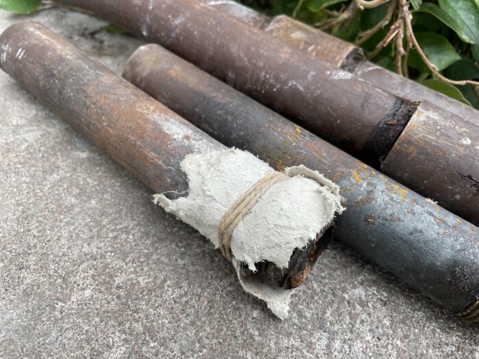 Gamla, rostiga rör lindade med trasig trasa på grov betongyta, med växtlighet i bakgrunden.