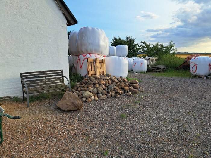 Lantgårdsmiljö med plastbalpressad hö, en bänk, stenar och prydnader vid en vit vägg.