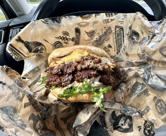 Oöppnad hamburgare i bil, smält ost, kött, grönsaker, omslagspapper med tryck, i förarsätet.