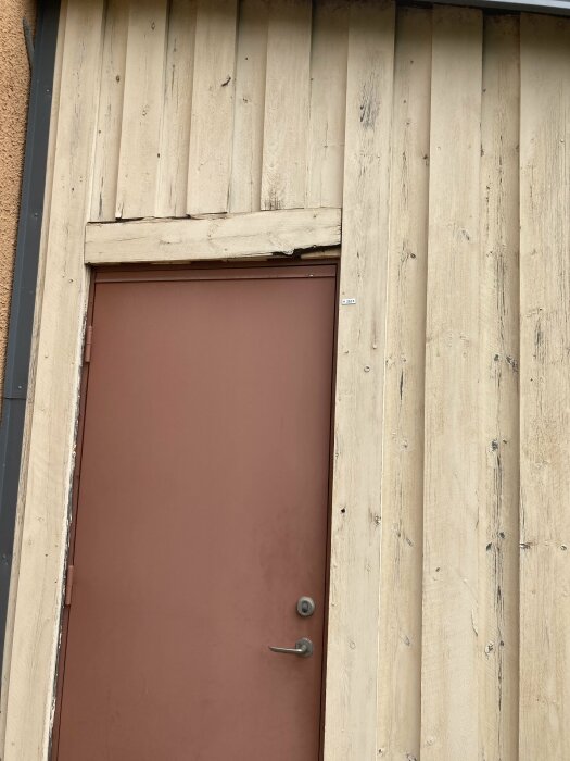 Brun dörr i en vertikal träpanelvägg, handled och lås synliga, slitna ytor.