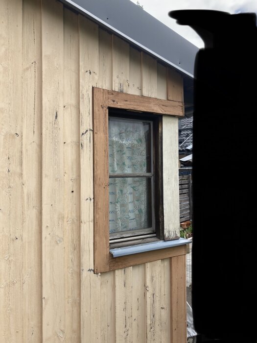 Fönster i sliten träram på ett trähus med mönstrad gardin och en mörk silhuett till vänster.
