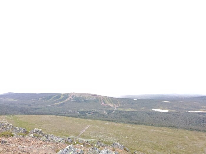 Öde fjällandskap med grå himmel, vegetation, några små vattenområden och grusiga vägar eller stigar.