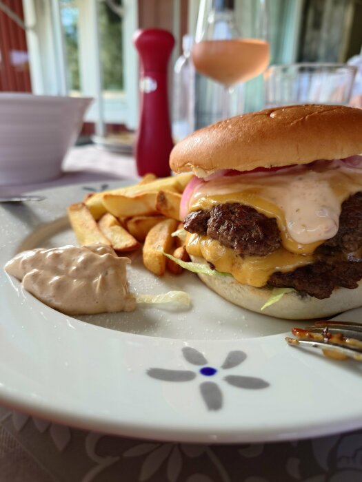 Cheeseburgare med pommes frites och sås på tallrik, kryddkvarn och rosévin i bakgrunden, hemtlig middagsmiljö.