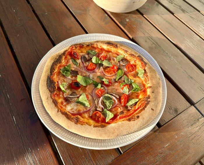 En pizza med tomat, basilika och svamp på ett träbord utomhus i solljus.