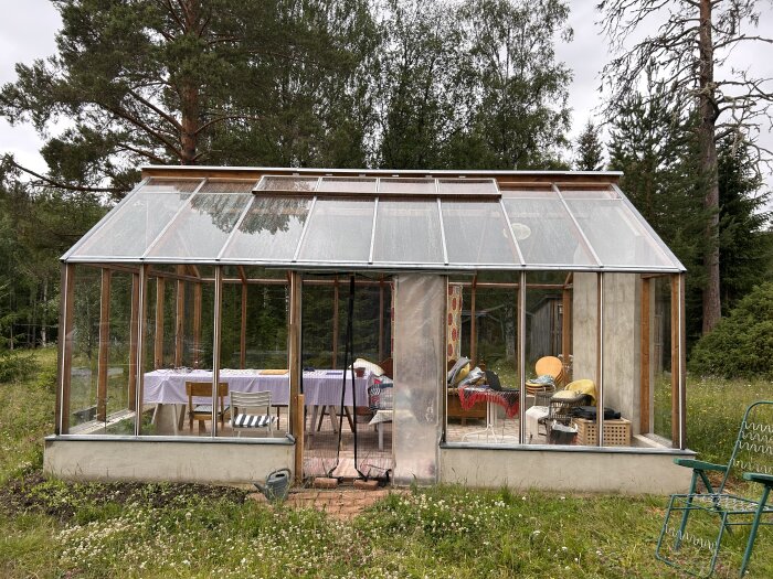 Växthus med möbler, trädgårdsredskap, skog i bakgrunden, molnig himmel, rustik atmosfär, betongfundament, transparent tak och väggar.