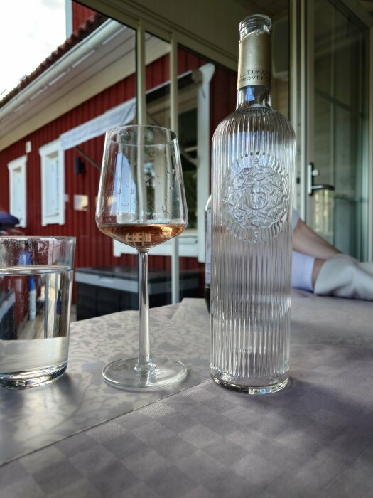 Vinflaska, glas med rosévin, vattenglas, checkrutig duk, veranda, röd stuga, avslappnad atmosfär.