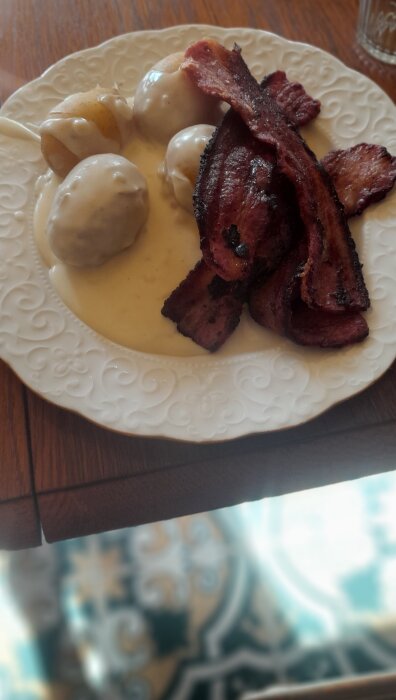 Köttbullar i gräddsås med krispig bacon på ett vitt, mönstrat fat.