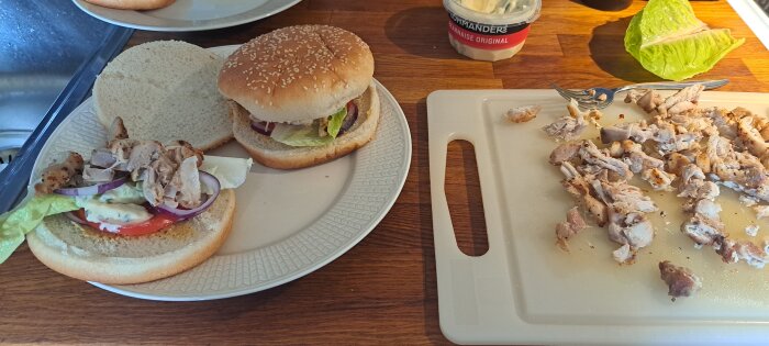 Två hamburgare med sallad och tomat, hackad kyckling på skärbräda, burk med bearnaisesås, träskiva.