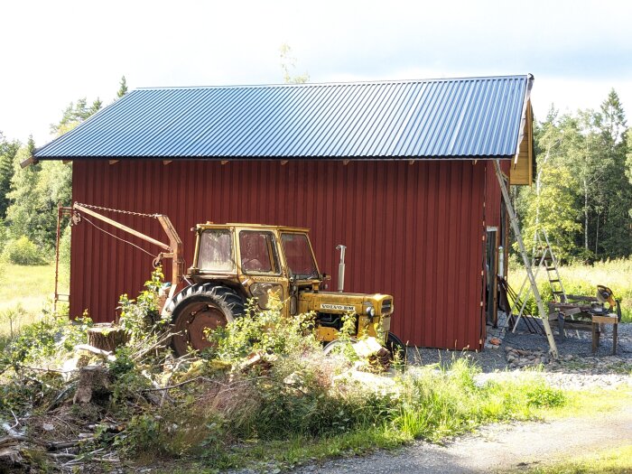 Röd lada, blått tak, gul traktor, grönska, avverkningsrester, soligt, lugn landsbygdsmiljö.